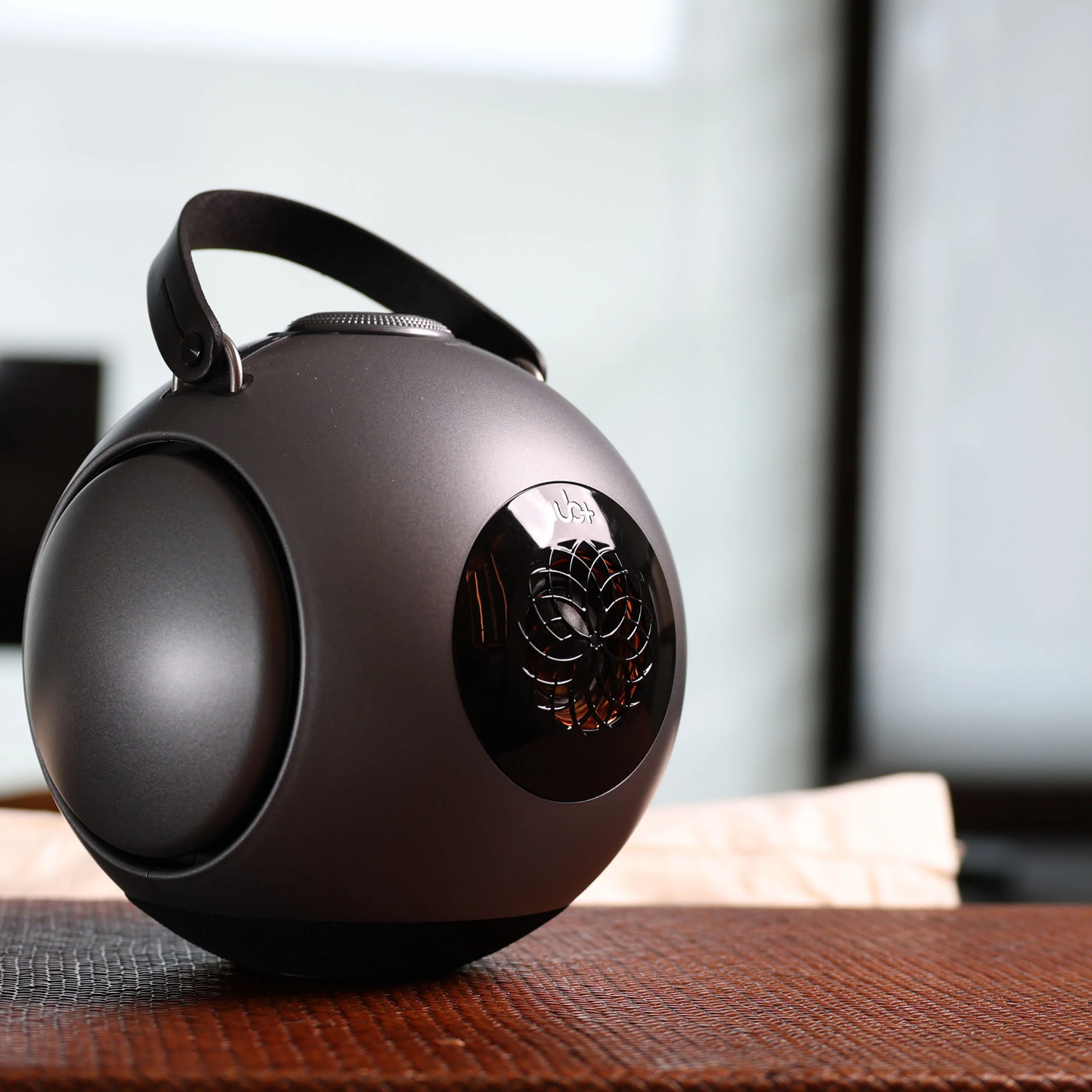 UB+ DB1 Doublebass portable speaker in metallic grey with black handle, displayed on a textured brown surface in a softly lit indoor setting.