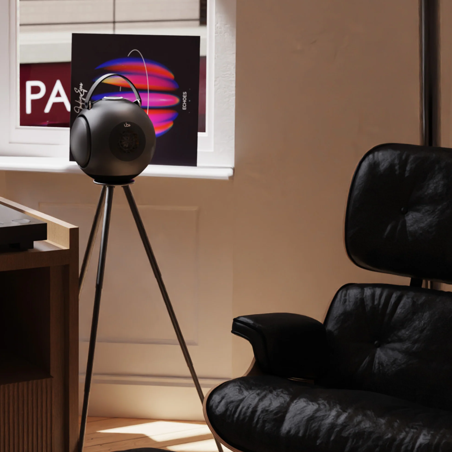 UB+ DB1 Doublebass portable speaker in metallic grey placed on a tripod stand in a modern room, next to a black leather chair and window.