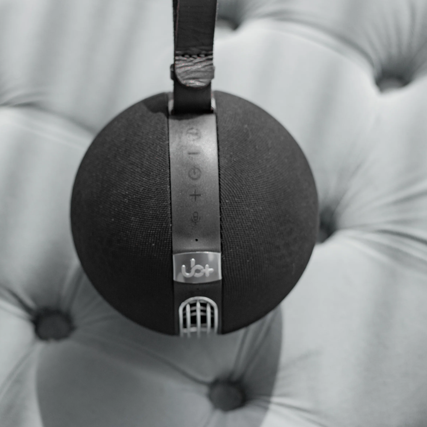 UB+ S1 Circle Bluetooth speaker in black, shown upright with a leather strap and silver top controls, resting on a soft tufted fabric surface.