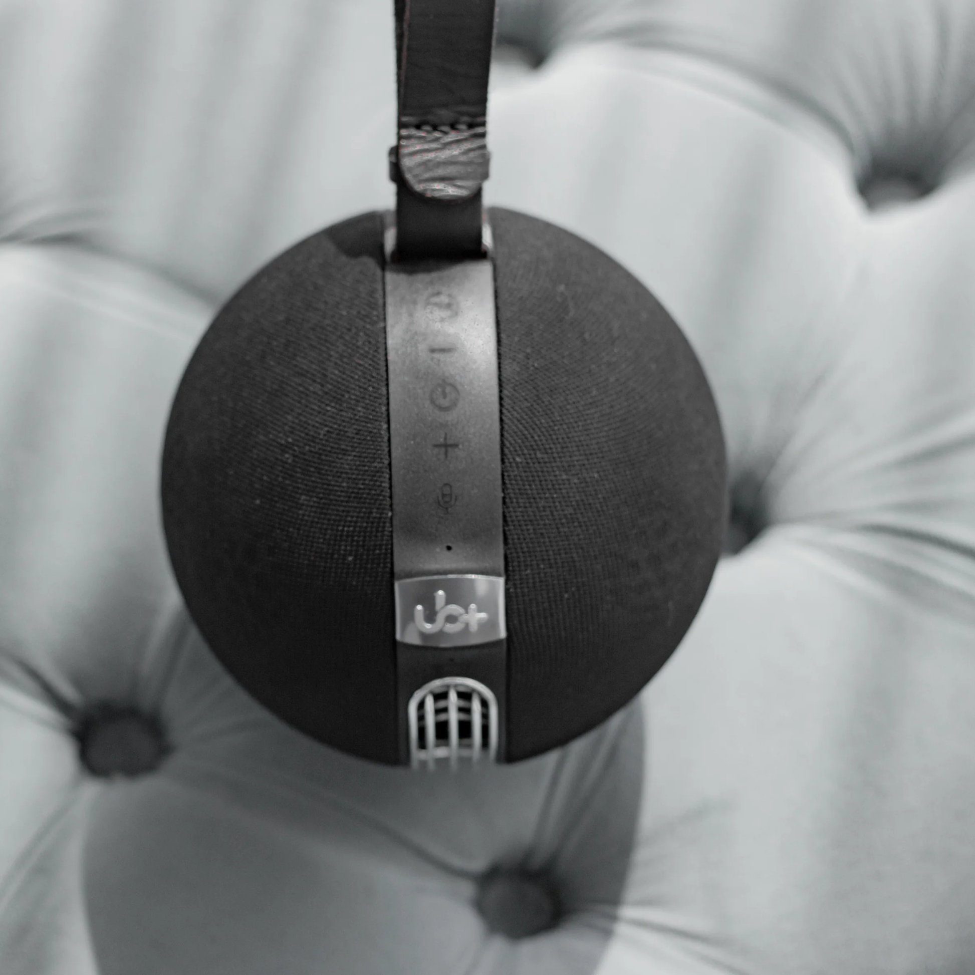 UB+ S1 Circle Bluetooth speaker in black, shown upright with a leather strap and silver top controls, resting on a soft tufted fabric surface.