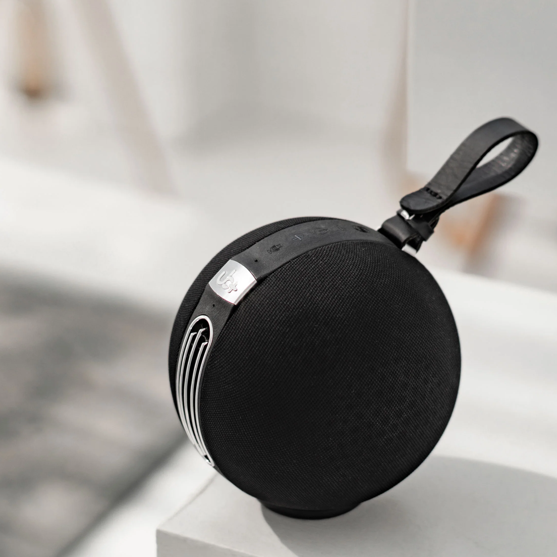 UB+ S1 Circle Bluetooth speaker in black, displayed on a white surface in a minimalist room with a leather strap and silver control panel visible.