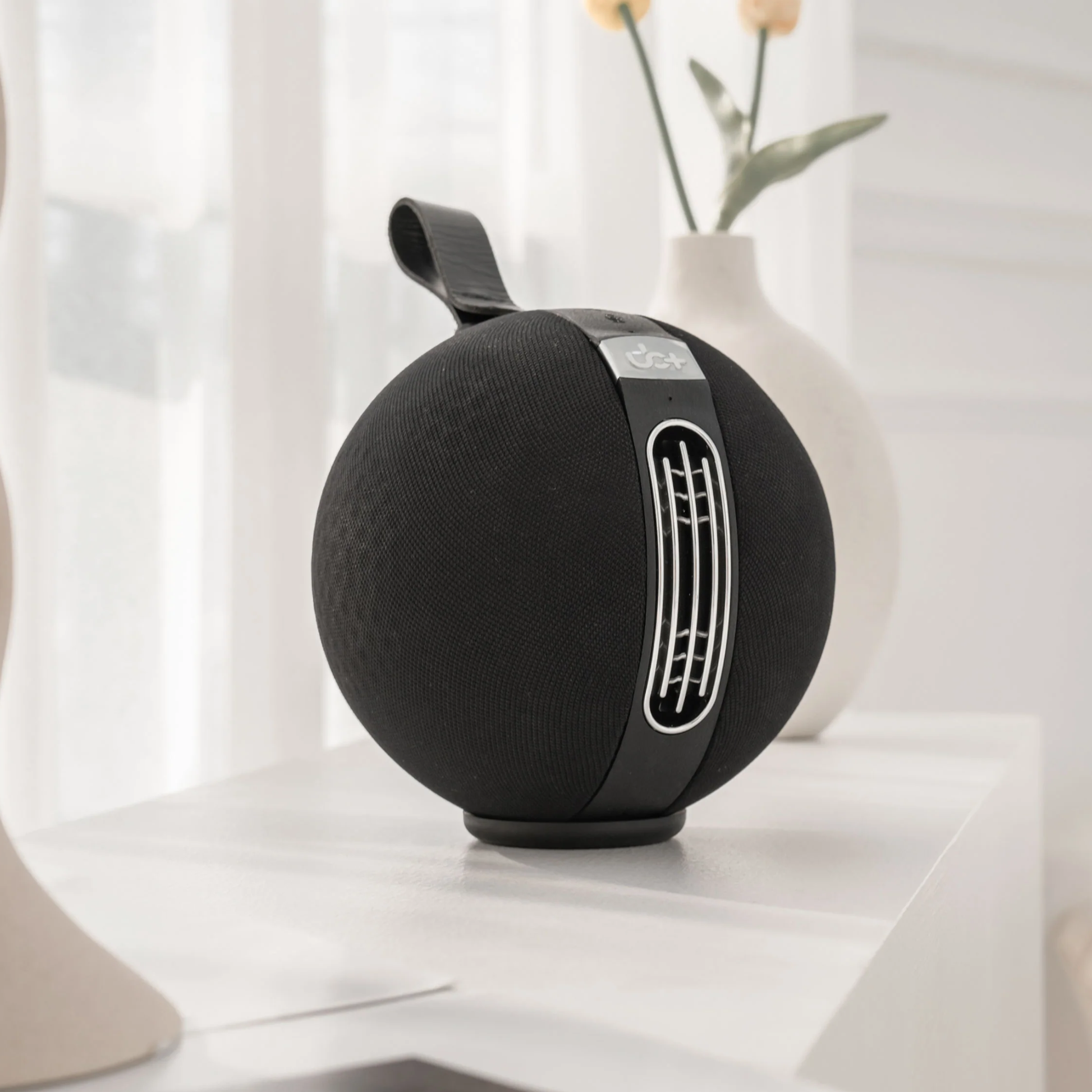 UB+ S1 Circle Bluetooth speaker in sleek black, angled on a white table in a bright modern room with decorative vases in the background.