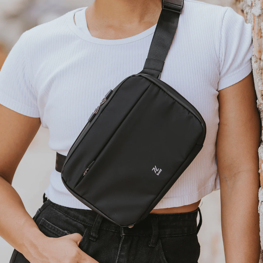 Woman wearing a black crossbody sling bag with adjustable strap over a white ribbed crop top and black jeans.
