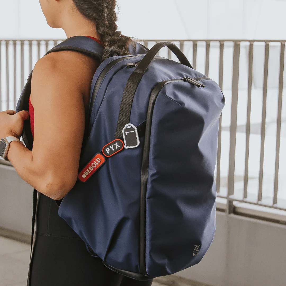 Woman with braided hair wearing a large navy blue backpack with #BeBold and PYX tags, standing outdoors near metal railings.