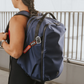 Woman with braided hair wearing a large navy blue backpack with #BeBold and PYX tags, standing outdoors near metal railings.