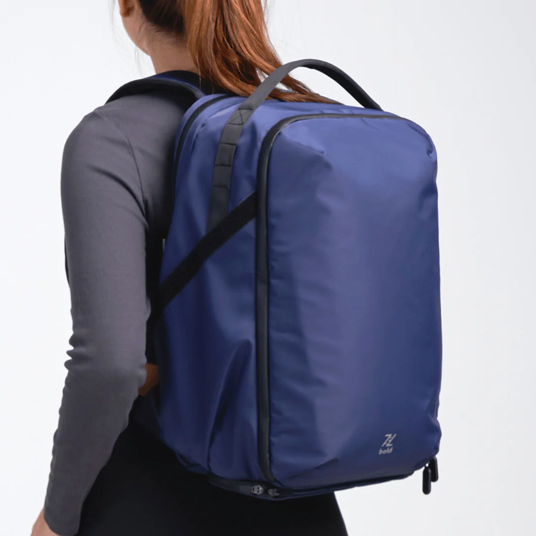 Woman wearing a large navy blue travel backpack with black straps and top handle, side view against a plain background.