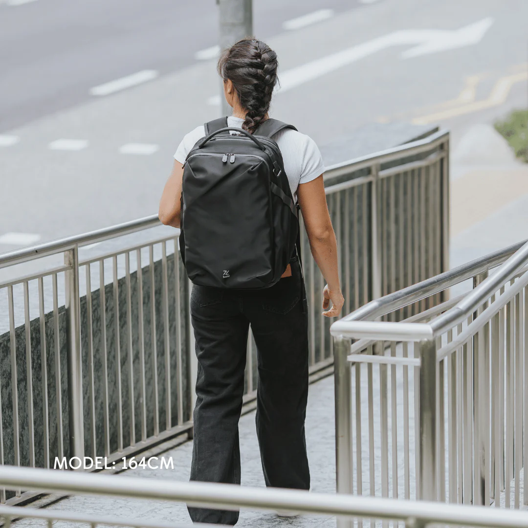 Woman with braided hair wearing a black backpack and white t-shirt, walking outdoors on a city ramp with railings, model height 164cm.